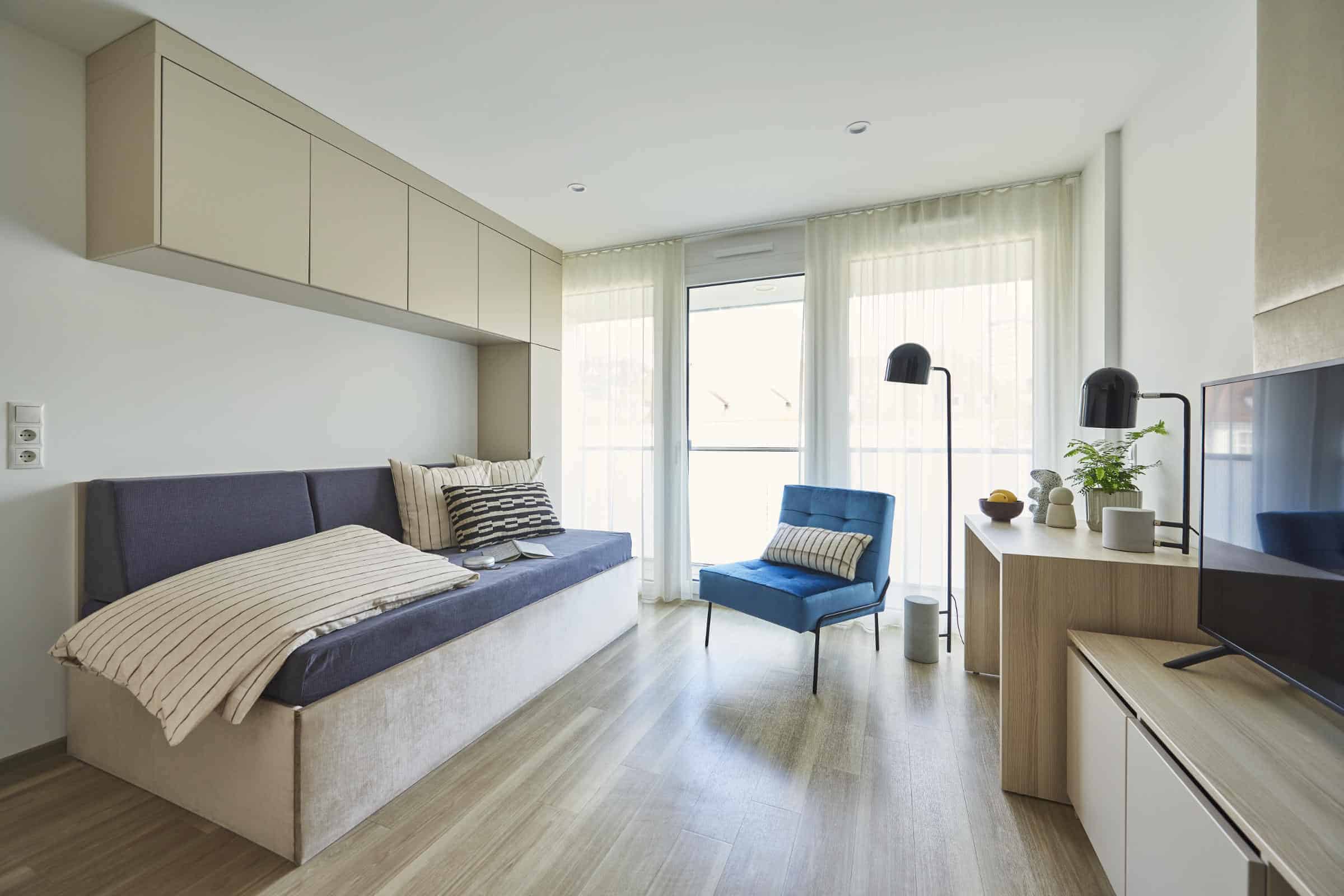 Apartment im Roomments Basic mit Tagesbett, blauem Sessel und Fensterfront